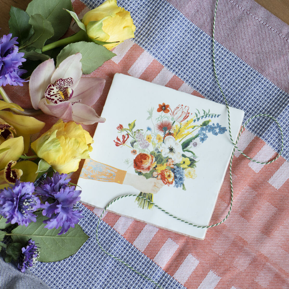Eine Keramikfliese mit einem bunten Blumenstrauß-Muster liegt auf einem gestreiften Stofftuch. Um die Fliese sind frische Blumen wie Orchideen und gelbe Tulpen dekoriert.