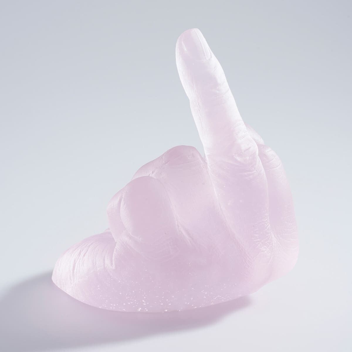 Eine Skulptur aus rosa transparentem Glas zeigt eine Hand, die den Mittelfinger hebt. Die Skulptur ist ein Kunstwerk von Ai Weiwei namens »Study of Perspective in Glass«. Sie liegt auf einer weißen, blanken Oberfläche.