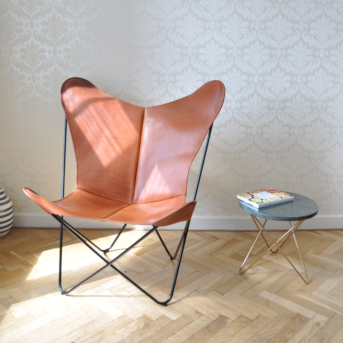 Ein eleganter Butterfly Chair aus braunem Leder auf einem metallenen Rahmen steht neben einem kleinen runden Beistelltisch mit goldenen Beinen und einer Tischplatte aus Glas. Der Raum hat helle Wände mit dezentem Ornamentmuster und einen Holzboden mit Fischgrätmuster. Auf dem Tisch liegen ein paar Bücher.
