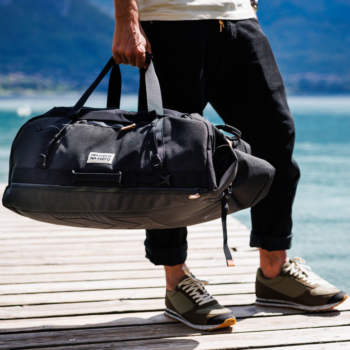 Eine Person trägt die schwarze Duffle »Smögen«. Die Tasche hat einen stabilen Griff, ist robust und geräumig. Im Hintergrund ist ein See und Berge zu sehen, und die Person steht auf einem hölzernen Steg.