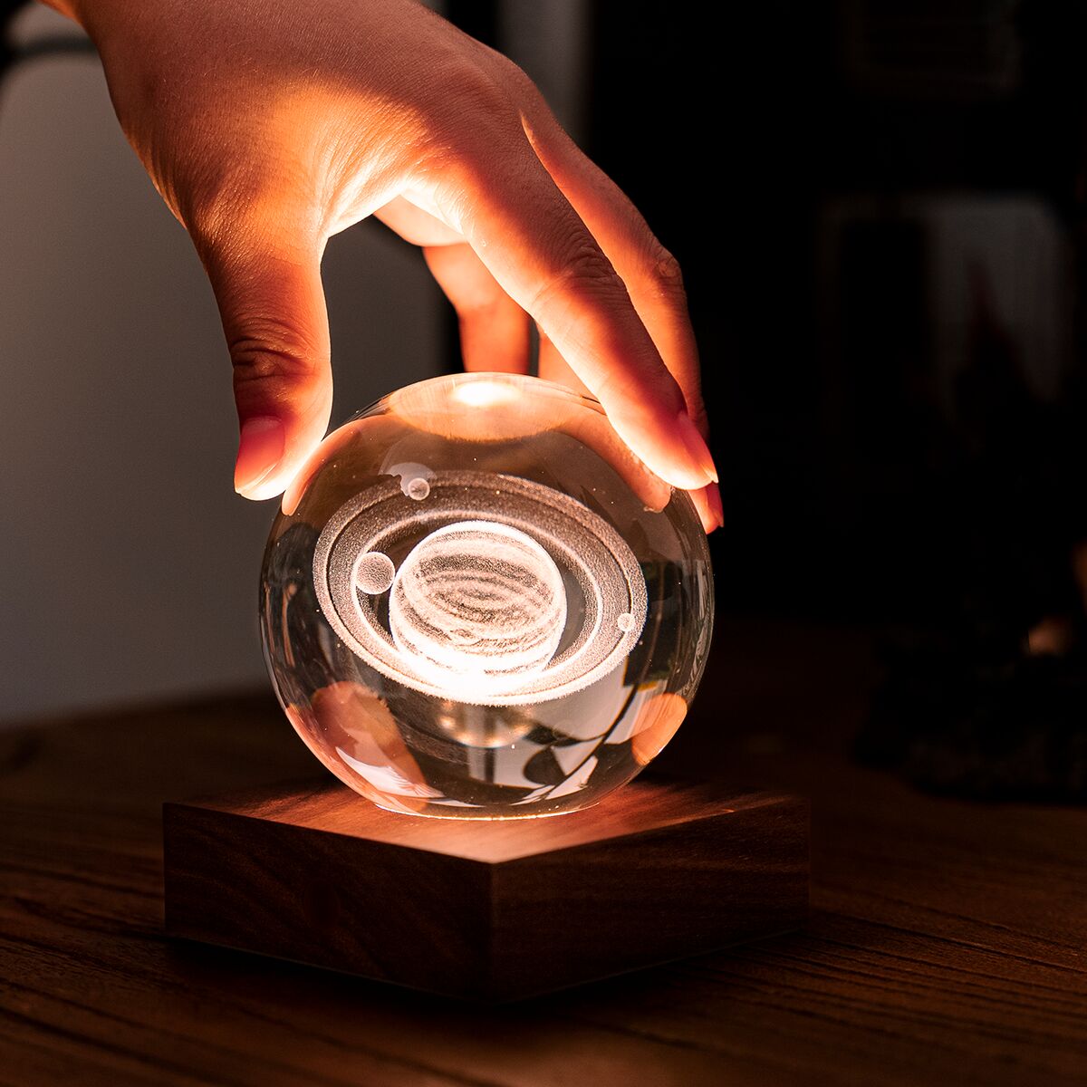 Eine dekorative Lampe namens »Amber Light« in Kugelform auf einem quadratischen Holzsockel. Die Kugel leuchtet von innen in warmem bernsteinfarbenem Licht. Eine Hand schwebt knapp über der Kugel. Die Lampe steht auf einer dunklen Oberfläche in einem modernen Wohnambiente.