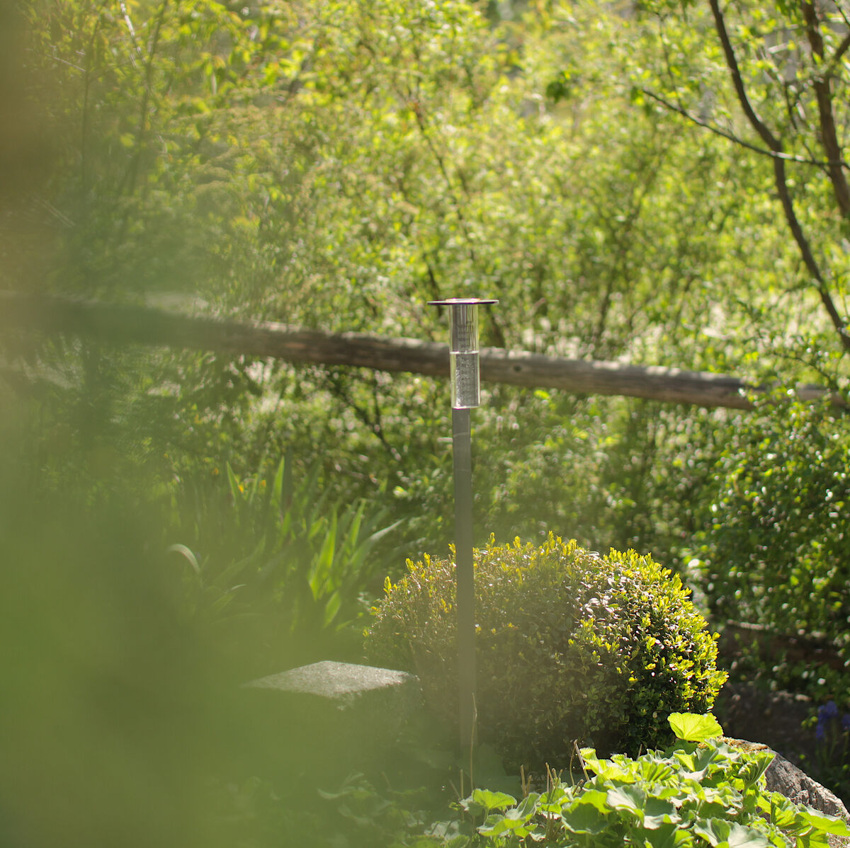 Eine Messstation im Garten, umgeben von üppigem Grün und Pflanzen. Es ist eine ruhige, naturnahe Szene mit Sonnenlicht, das durch die Bäume scheint.