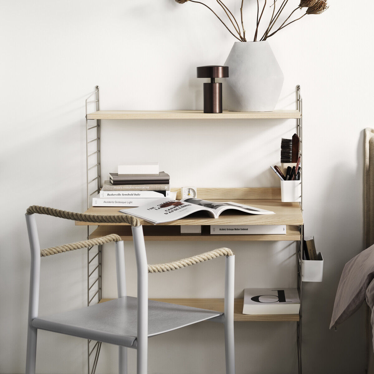 Ein minimalistischer Arbeitsplatz bestehend aus einem schlanken, modularen String-Möbelsystem mit Regalen, einer Arbeitsfläche und einer Metallstuhl mit einem Kissen. Der Schreibtisch ist in einem modernen, schlichten Design, das perfekt zu einem hellen, gut organisierten Raum passt. Neben dem Arbeitsplatz befindet sich ein schlichtes Bett. Auf dem Schreibtisch stehen Bücher, Schreibutensilien und eine Vase mit getrockneten Blumen. Die Farben im Bild sind Weiß, Beige, Grautöne und verschiedene Holztöne.