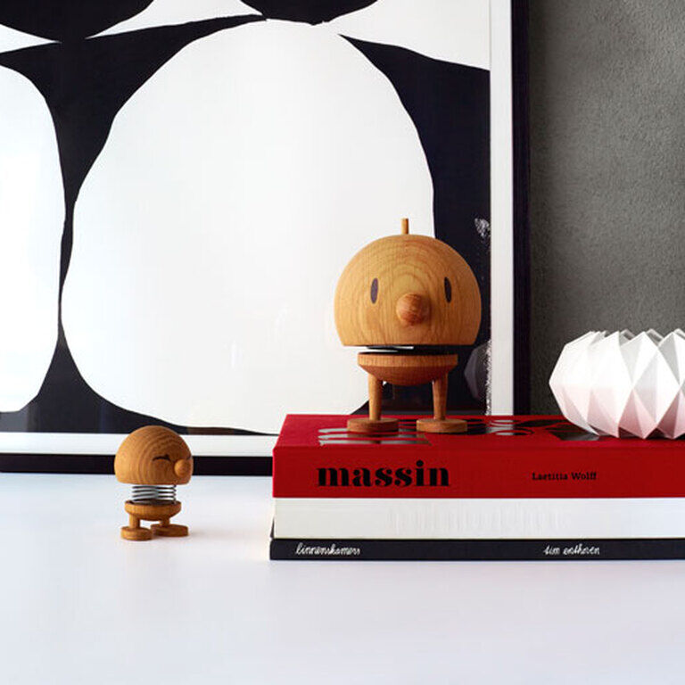 Das Bild zeigt zwei Hoptimist Bumble Figuren aus Eiche auf einem weißen Hintergrund. Eine Figur steht auf einem Stapel von Büchern, und im Hintergrund ist ein minimalistisches Kunstwerk mit schwarzen und weißen Formen zu sehen.