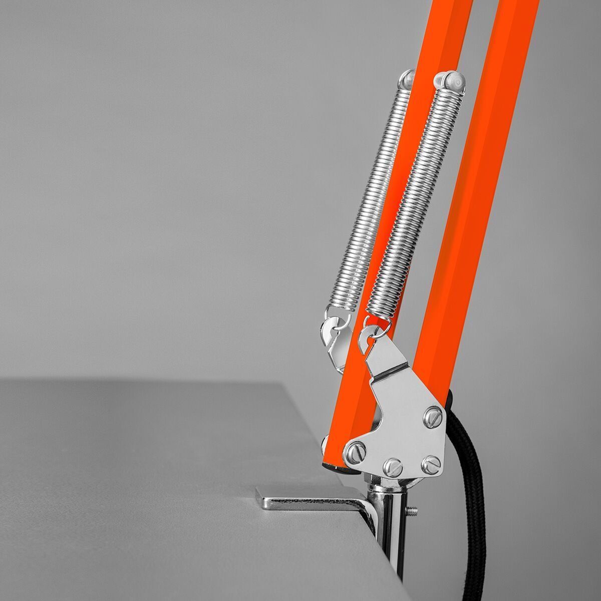 Eine Tischleuchte »Federzug« mit Klemmfuß aus der ZEIT-Sonderedition, die an einer grauen Oberfläche befestigt ist. Die Lampe verfügt über eine orangefarbene, verstellbare Halterung mit sichtbaren Federn und einem stabilen Klemmsockel.