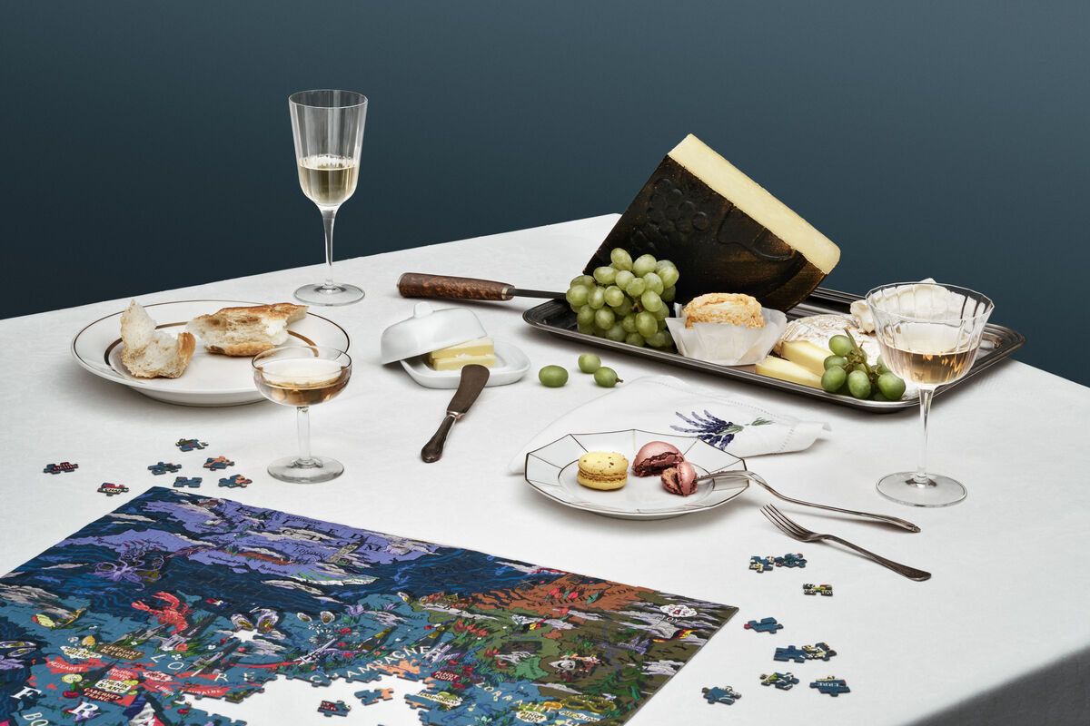 Auf einem elegant gedeckten Tisch liegt ein fast fertiggestelltes Puzzle mit einem Bild von Frankreich. Daneben stehen Weingläser mit Weißwein, ein Teller mit Käse, Trauben und Brotscheiben. Die Farben des Tisches und der Gegenstände sind überwiegend weiß, grün, gold, blau und lila.