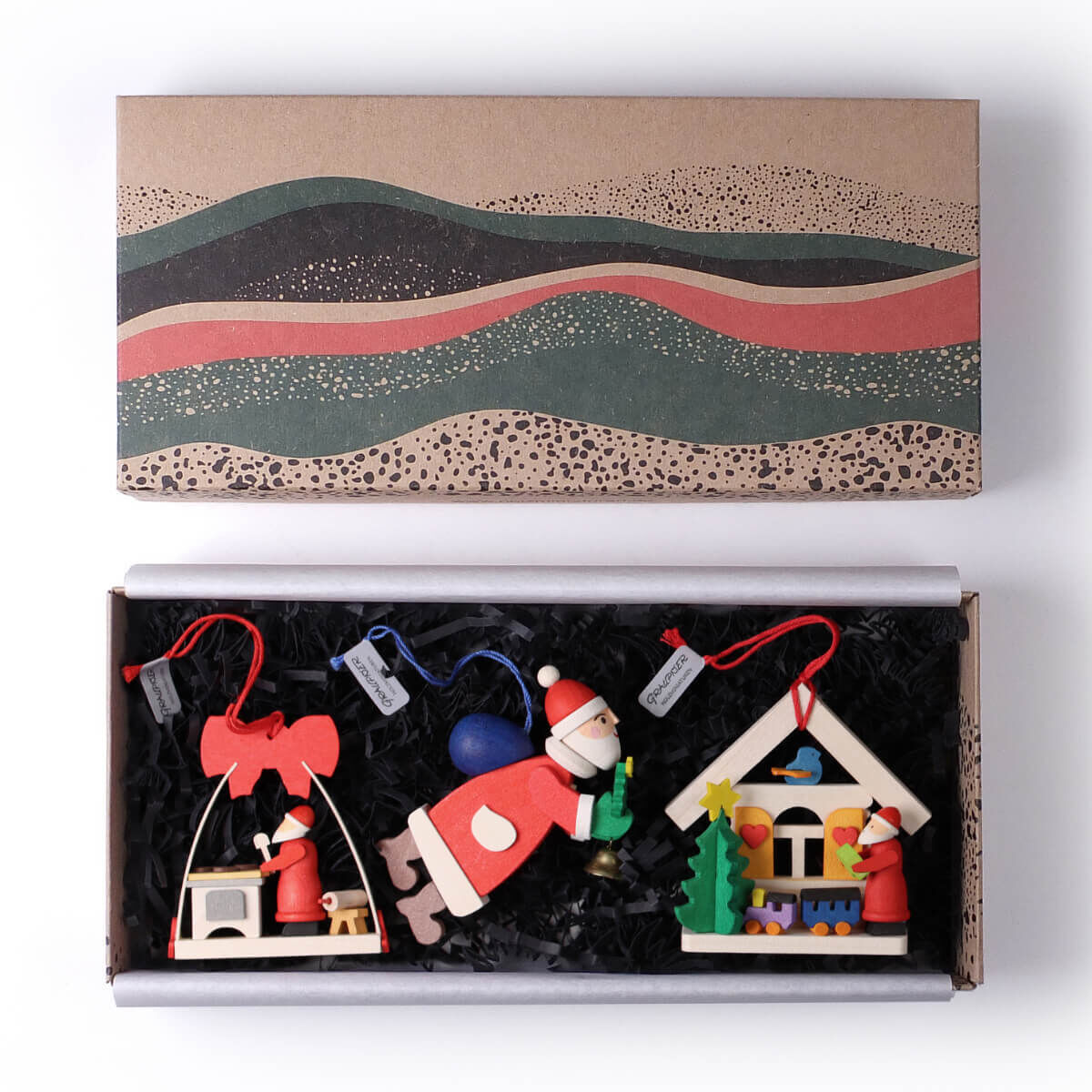 Dreiteiliges Geschenk-Set mit Weihnachtsbaumschmuck bestehend aus handgefertigten Holzfiguren in Form von Weihnachtsmännern, farbenfroh gestaltet in den Farben Rot, Weiß, Blau und Grün. Verpackt in einer dekorativen Box.