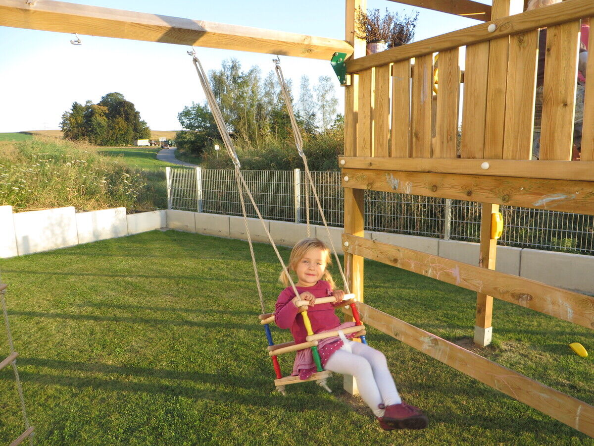 Ein kleines Mädchen sitzt auf einer Schaukel aus Buchenholz, die an einem hölzernen Spielgerüst im Garten befestigt ist. Im Hintergrund sind Gras, ein Zaun und Bäume zu sehen. Das Kind trägt ein rotes Oberteil und weiße Strumpfhosen.