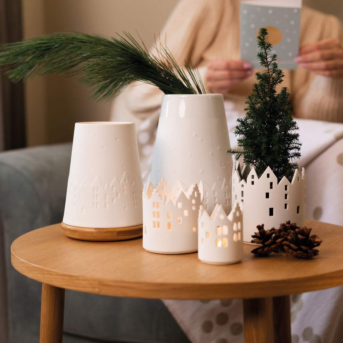 Weißes Windlicht-Set »Stadtlicht« auf einem Holztisch, verziert mit Tannenzweigen und einem kleinen Weihnachtsbaum, im gemütlichen Wohnzimmer-Ambiente.