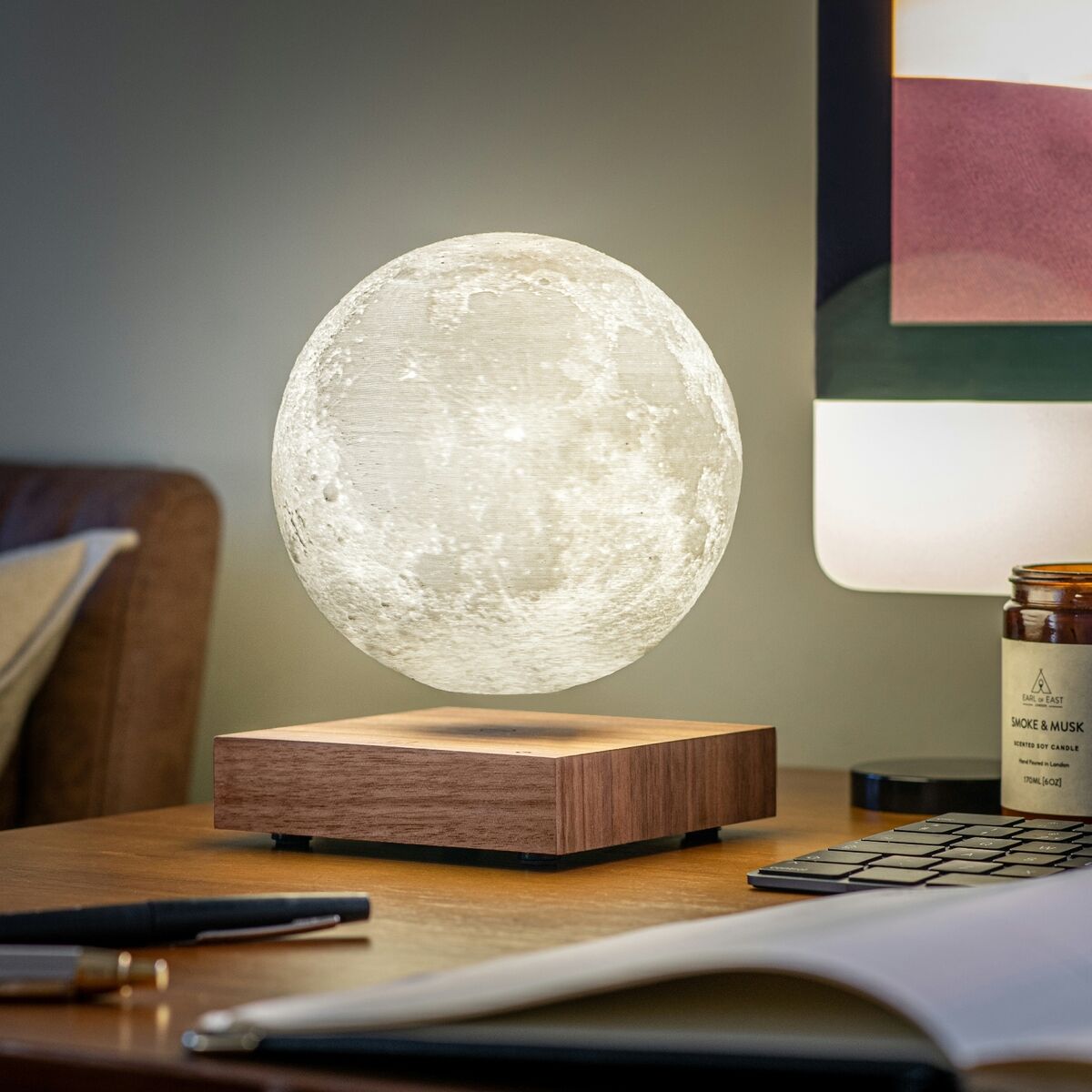 Eine schwebende Smart Moon Lampe mit einer realistischen Mondoberfläche, die über einer rechteckigen Holzbasis leuchtet. Die Lampe ist auf einem Schreibtisch neben einem Notizbuch und einer Lampe mit braunem Schirm platziert.