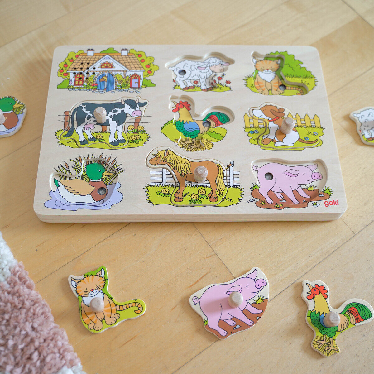Auf dem Bild ist ein Holz-Soundpuzzle mit Bauerhofmotiven zu sehen. Das Puzzle zeigt verschiedene Bauernhoftiere wie Katze, Schwein, Hahn, Pferd, Schaf, Ziege und Ente. Einige Puzzleteile liegen außerhalb des Puzzles auf einem hellen Holzboden, daneben ist ein Teil eines Teppichs sichtbar. Die Tiere sind farbig illustriert und kinderfreundlich gestaltet.