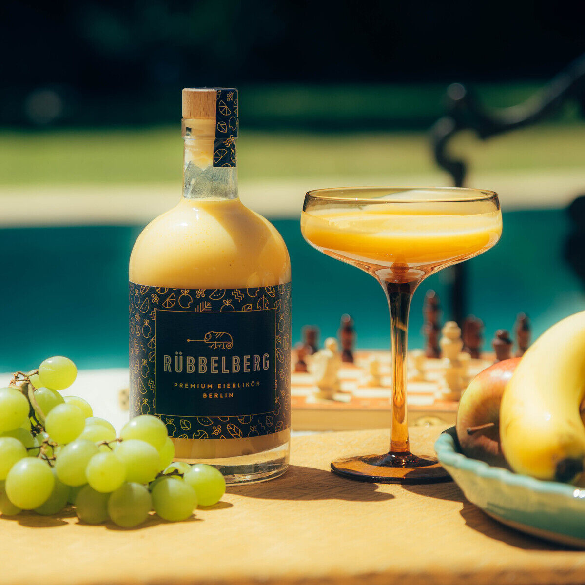 Eine stilvolle Flasche »Premium Eierlikör« von Rübbelberg steht neben einem eleganten Likörglas, das mit Eierlikör gefüllt ist. Im Vordergrund liegen frische grüne Weintrauben und Bananen auf einem Holzbrett. Im Hintergrund ein unscharfer Pool und Schachbrett, sonnige Atmosphäre.