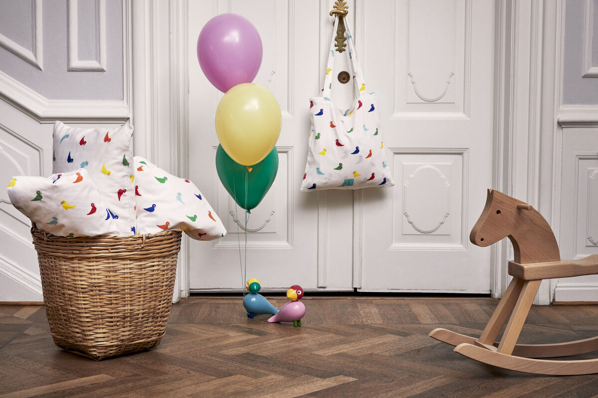 Ein Kinderzimmer mit einer Holzpferd-Wippe, einem geflochtenen Korb mit einer weißen Decke, die mit bunten Vogelmotiven bedruckt ist, bunten Luftballons und zwei farbenfrohen Holzvögeln 'Sunshine' von Kay Bojesen auf dem Holzboden. Im Hintergrund eine weiße Tür mit einer Dekotasche, ebenfalls mit bunten Vogelmotiven.