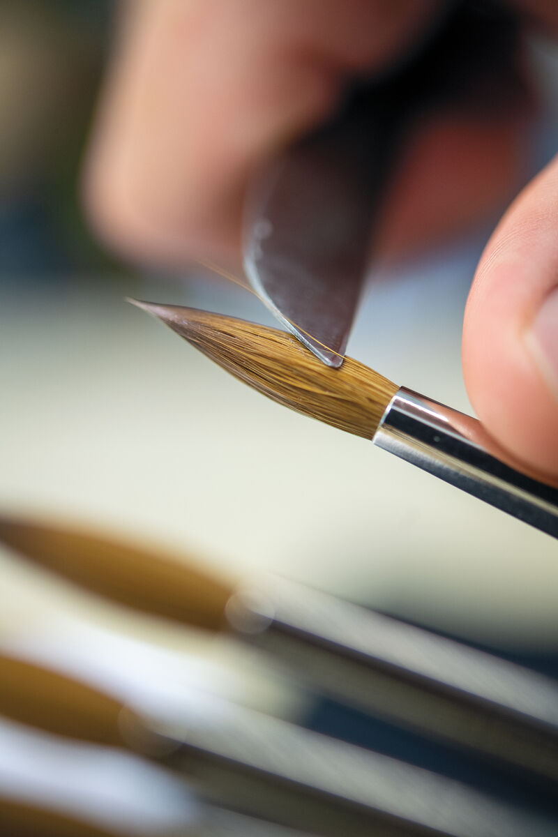 Nahaufnahme von einem traditionell gefertigten Aquarellpinsel mit einer Ethergraf®-Metallspitze, der in einer Hand gehalten wird. Im Hintergrund sind weitere Pinsel zu sehen.