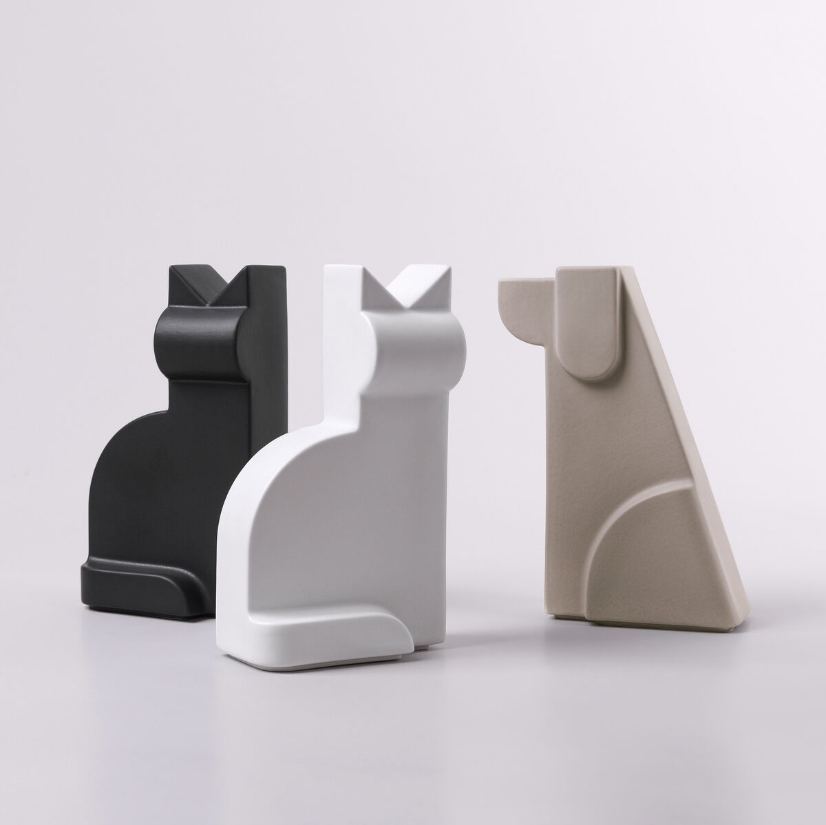 Drei minimalistische Porzellanfiguren in Form von Katzen in den Farben Schwarz, Weiß und Beige auf einem grauen Hintergrund. Jede Figur hat ein modernes und schlichtes Design.