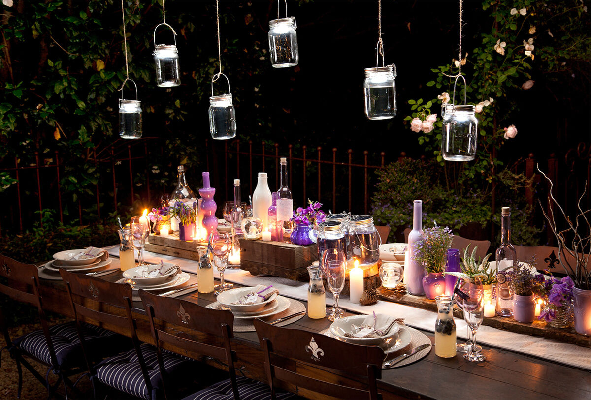 Ein festlich gedeckter Tisch im Freien bei Nacht, dekoriert mit hängenden Sonnenglas Classic Solarleuchten und Kerzen. Der Tisch ist mit Geschirrsets, lila und weißen Flaschen sowie verschiedenen Dekorationen geschmückt. Eine gemütliche und einladende Atmosphäre für eine Gartenparty.