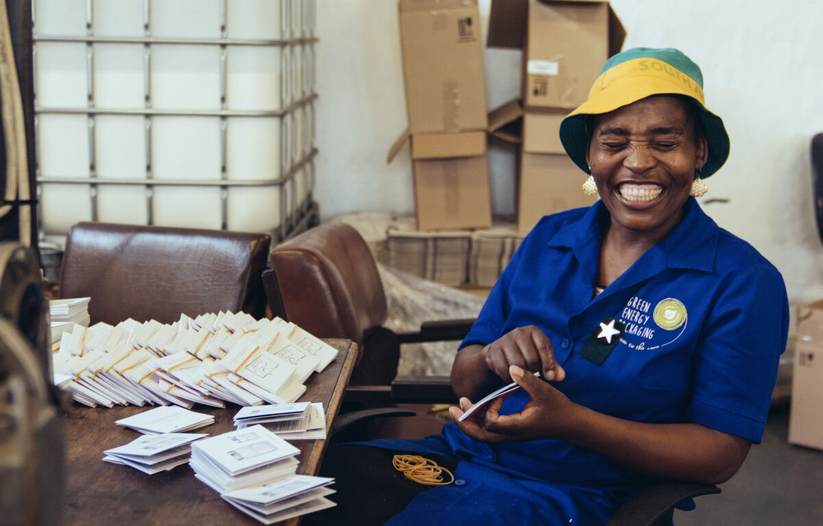 Eine lächelnde afrikanische Frau in blauer Arbeitskleidung bastelt im Werkstattbereich Glaslampen und Verpackungen. Auf ihrer Arbeitskleidung steht 'Africa Direct & Fair Trade'. Im Hintergrund sind Kartons und verschiedene Arbeitsmaterialien zu sehen.