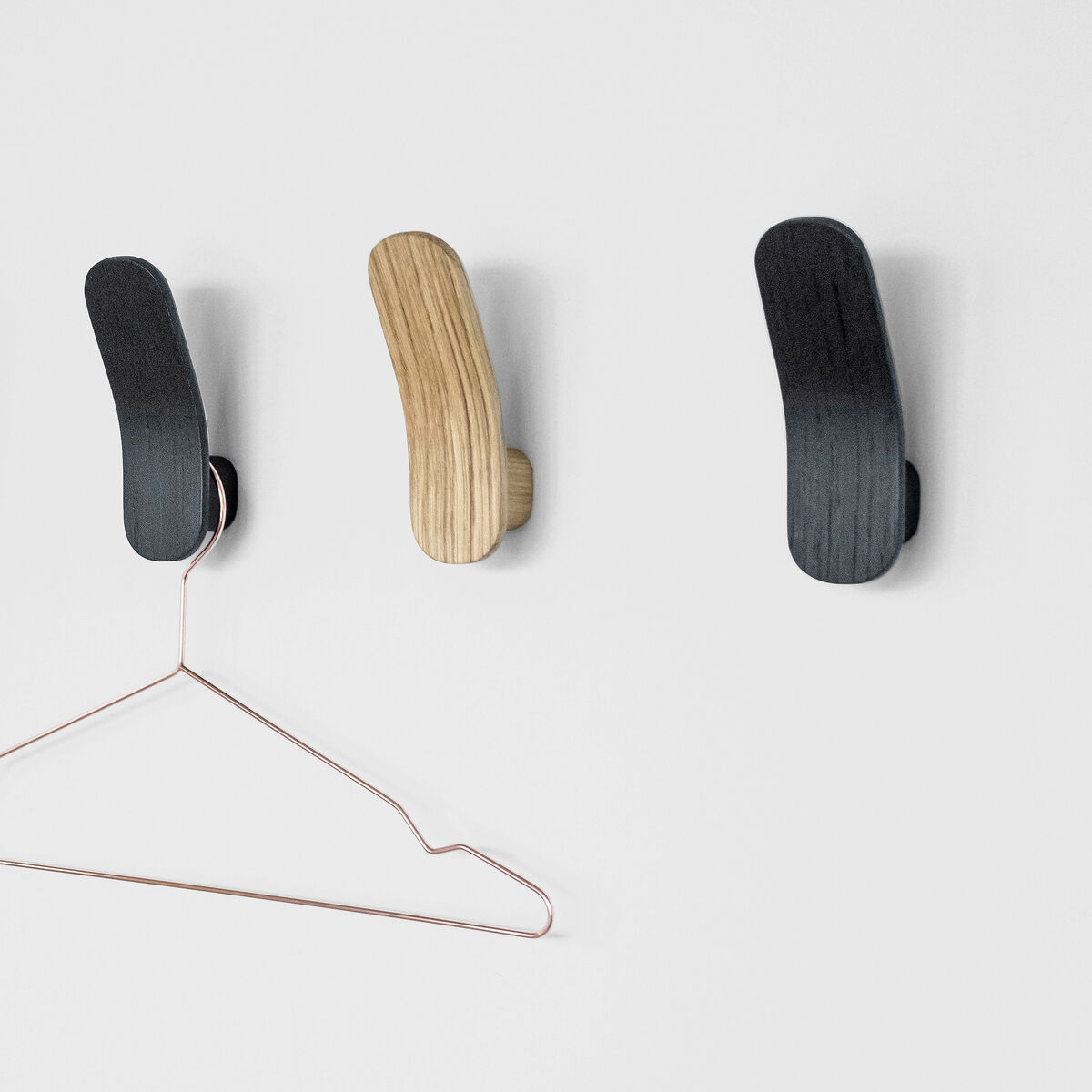 Vier Kleiderhaken aus Eiche, zwei in natürlicher Holzfarbe, zwei in schwarz angestrichen, befestigt an einer weißen Wand. Ein kupferfarbener Kleiderbügel hängt an einem der Haken.