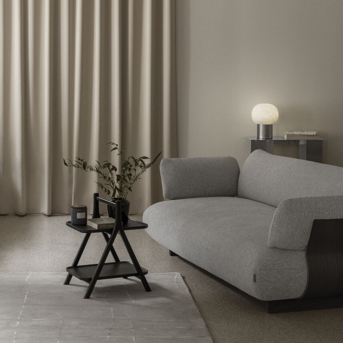 Modern eingerichtetes Wohnzimmer mit beigem Vorhang, grauem Teppich, heller Wand und Designer-Sofa. Im Zentrum steht der schwarze Beistelltisch "ETA" von blomus mit Dekozweigen, flankiert von dunklen Podesten, dekorativen Objekten und einer Leuchte auf Beistelltisch.