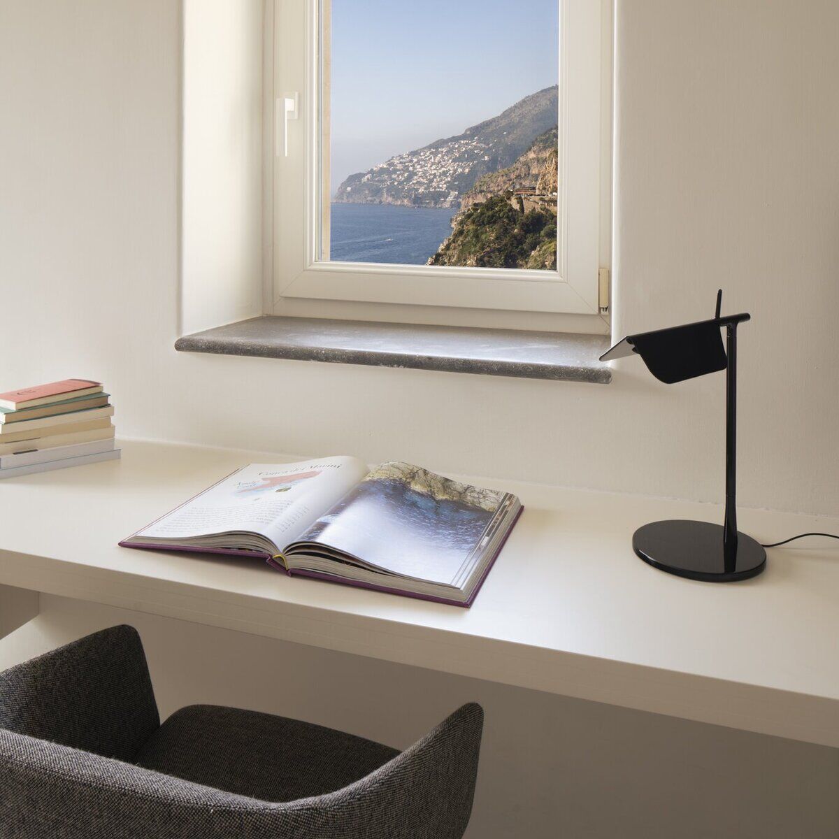 Ein moderner Schreibtisch mit einer geöffneten Zeitschrift, einem blick auf eine Küstenlandschaft aus einem Fenster und einer schwarzen Tischleuchte 'TAB' auf einem weißen Tisch. Designorientierter Arbeitsplatz mit funktionaler Beleuchtung.
