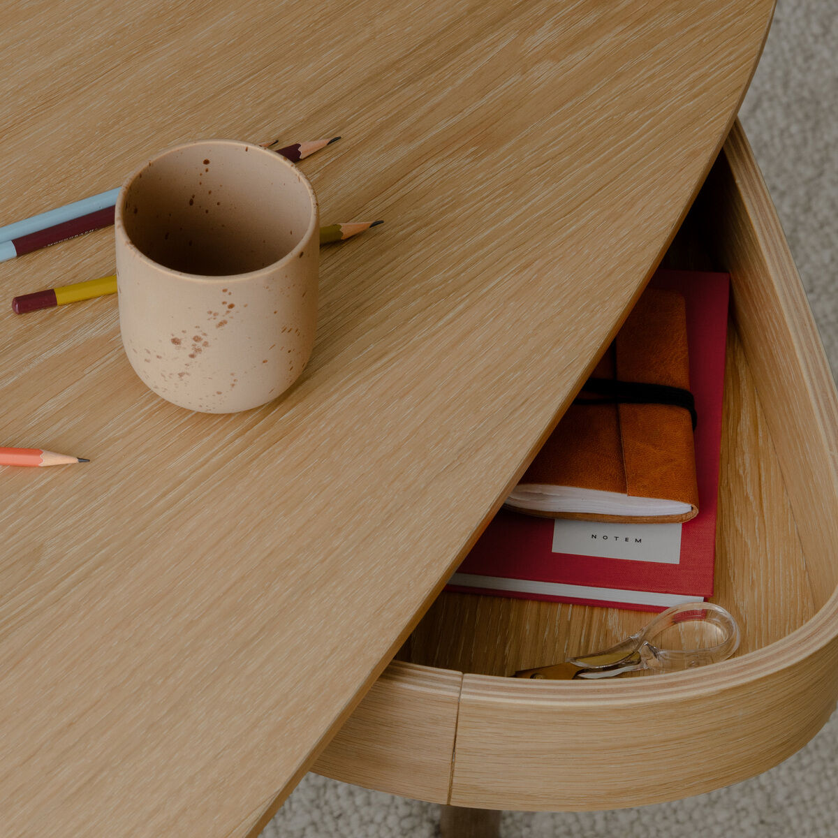 Ein moderner Schreibtisch aus hellem Holz mit organischer Form steht auf einem hellen Teppich. Auf der Oberfläche liegen Buntstifte und eine beige Tasse. Die leicht geöffnete Schublade enthält ein rotes Notizbuch, ein weiteres Buch, eine Brille und einen Stift.