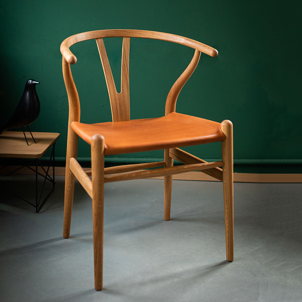 Die ZEIT Sonderedition »CH24« Wishbone Chair besteht aus Eichenholz mit einer Sitzfläche aus Leder. Der Stuhl hat ein minimalistisches und skandinavisches Design und steht vor einer grünen Wand und grauem Boden. Ein schwarzer Dekorvogel steht auf einem kleinen Tisch neben dem Stuhl.