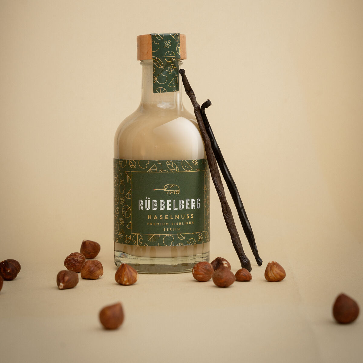 Eine kleine Glasflasche mit grünem Etikett der Marke Rübbelberg Haselnuss Eierlikör steht auf hellem Untergrund. Neben der Flasche liegen Haselnüsse und zwei Vanilleschoten, die eine hochwertige und natürliche Produktpräsentation vermitteln.