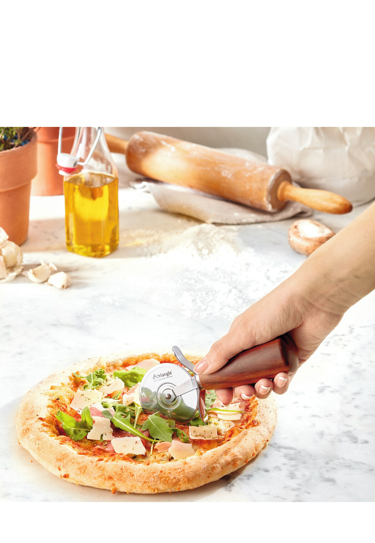 Eine Hand hält einen Pizzaschneider »Soul« aus Pflaumenholz über eine frisch gebackene Pizza. Im Hintergrund stehen Zutaten wie ein Olivenölflasche, Kräuter und ein Nudelholz auf einem weißen Marmortisch.