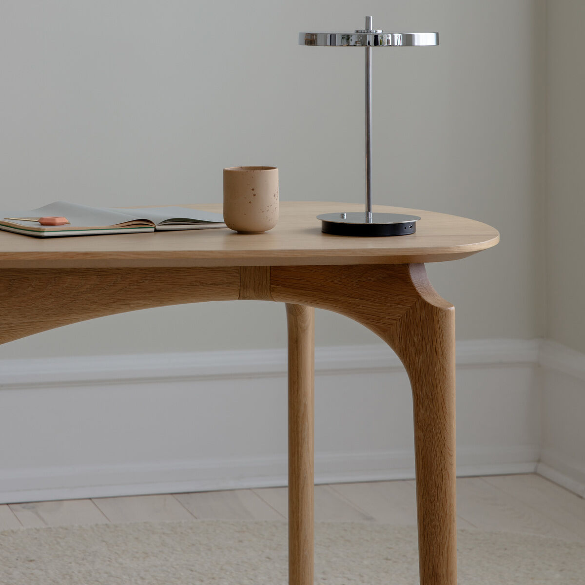 Ein moderner, minimalistisch gehaltener Schreibtisch aus hellem Holz steht vor einer neutralen Wand. Auf dem Tisch liegen ein geöffnetes Magazin, eine stilvolle Schreibtischlampe mit Metallakzenten und eine beige Tasse. Der Raum wirkt aufgeräumt und hell, ideal als Arbeitsplatz oder für kreative Tätigkeiten.