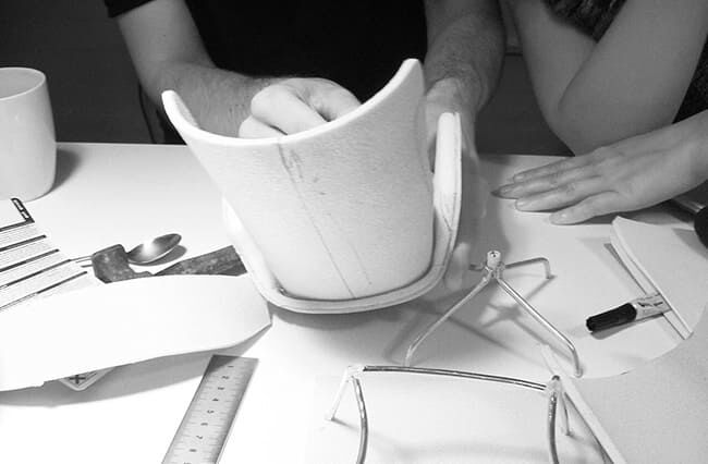 Zwei Hände, die an einem Designprojekt arbeiten, konzentrieren sich auf einen weißen Prototyp und sind von verschiedenen Werkzeugen und Materialien auf einem Tisch umgeben.