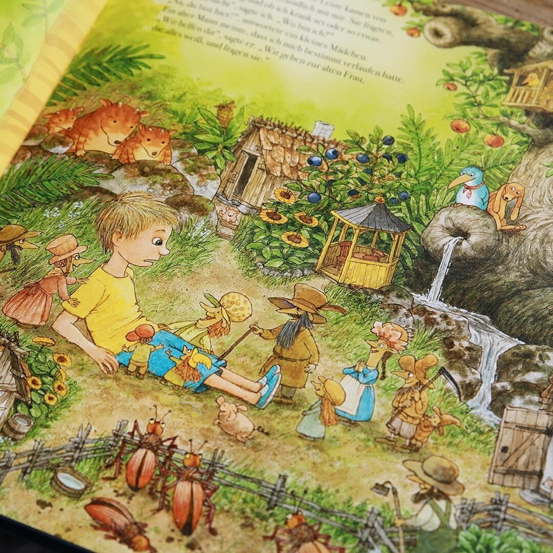 Ein illustriertes Kinderbuchbild aus 'Der Weg nach Hause' zeigt eine farbenfrohe Waldszene mit einem kleinen Jungen in gelber Kleidung, umgeben von magischen Waldwesen, kleinen Häusern und Pflanzen. Im Hintergrund ist ein kleiner Wasserfall und ein großer Baum mit einem Baumhaus zu sehen.