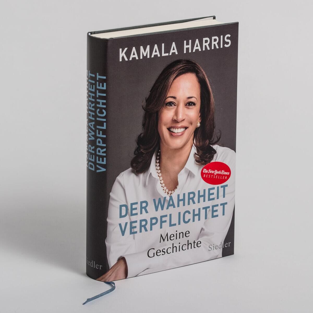 Das Bild zeigt das Buch 'Der Wahrheit verpflichtet: Meine Geschichte' von Kamala Harris. Es ist ein Hardcover mit einem grauen Hintergrund und einem Bild von Kamala Harris, die ein weißes Hemd trägt. Rechts befindet sich ein runder roter Aufkleber mit der Aufschrift 'New York Times Bestseller'. Der Titel des Buches und der Autorname sind in weißer und blauer Schrift geschrieben. Ein blaues Leseband ist ebenfalls sichtbar.