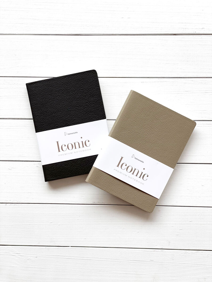 Zwei Notizbücher »Iconic« im DIN A5 Format auf einem weißen Holzuntergrund. Ein Notizbuch ist schwarz und das andere beige, beide werden von einer weißen Banderole umgeben.