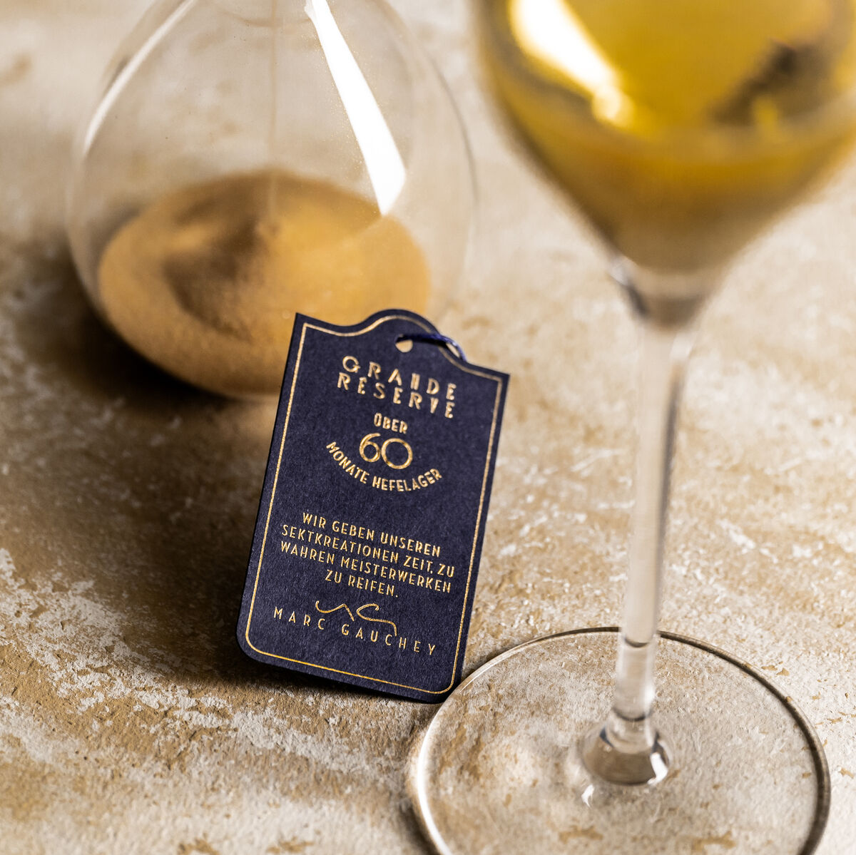 Edle Flasche von Geldermann Grande Réserve neben Riedel Glas auf einem eleganten Tisch; Hintergrund aus beigem Material und Sanduhr-Element.