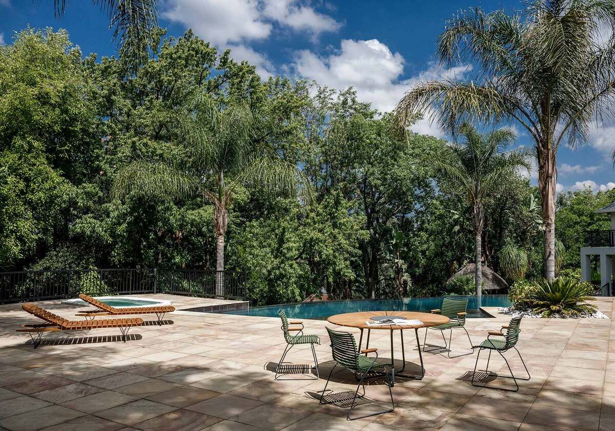 Ein Außenbereich mit grünem Laub, einem Pool und modernen Gartenmöbeln, darunter Liegen und ein runder Tisch mit Sesseln.