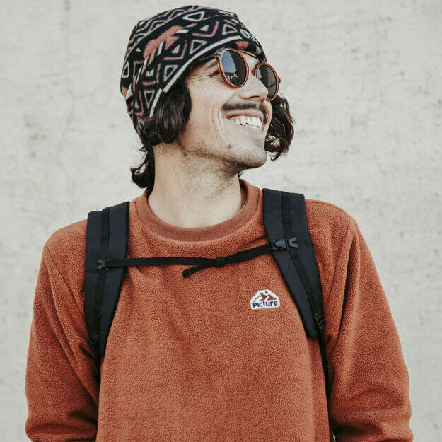 Eine Person mit einer rotbraunen Fleecejacke, schwarzem Rucksack, gemusterter Mütze und Sonnenbrille steht vor einer beigen Wand und lächelt.