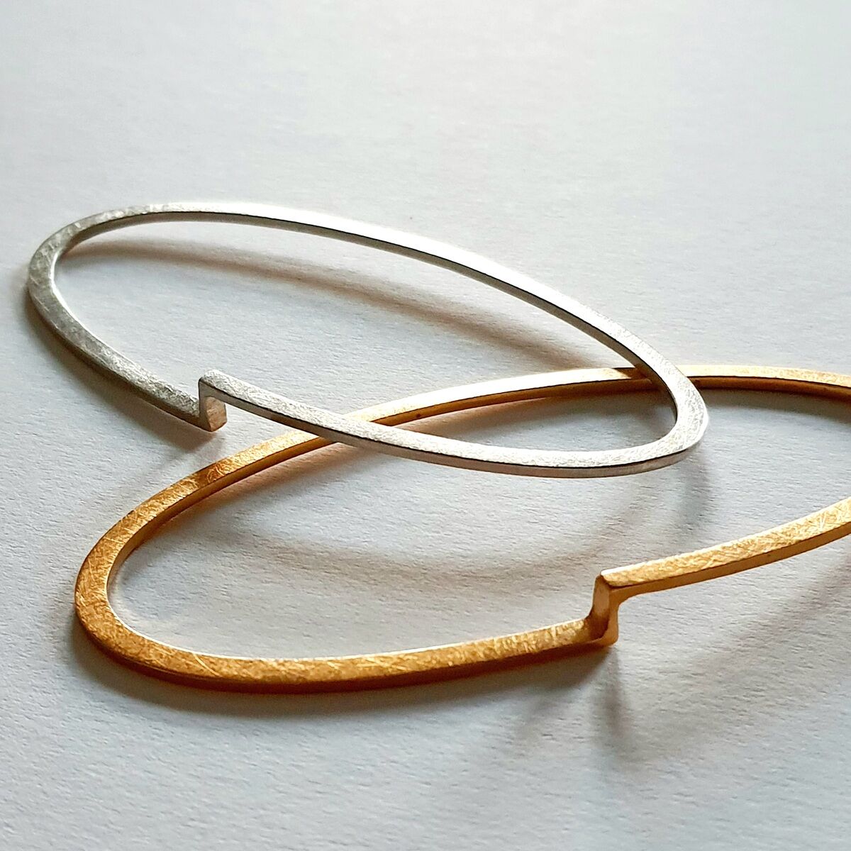 Zwei schlichte, moderne Armreifen, einer in Gold und einer in Silber, liegen überkreuzt auf einer weißen Oberfläche.