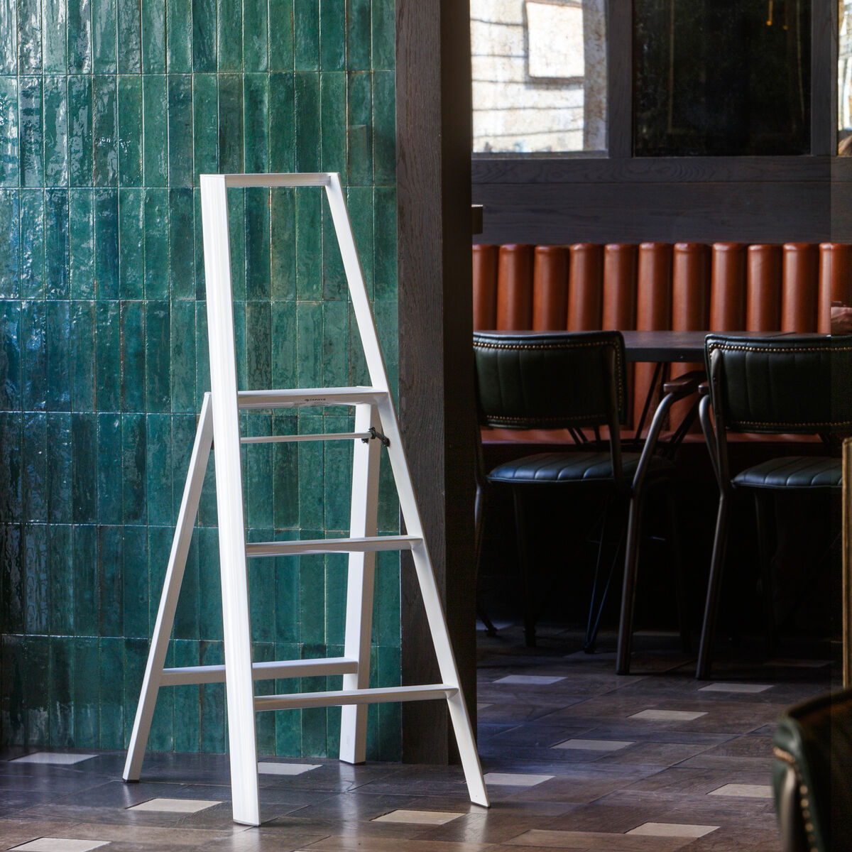 Weiße Lucano Trittleiter mit drei Stufen steht in einem modernen Innenraum neben einer grünen gefliesten Wand und einem Essbereich mit dunklen Stühlen und Tischen. Das Foto betont das elegante Design und die Funktionalität der Leiter, geeignet für Haushalts- oder Arbeitsräume.