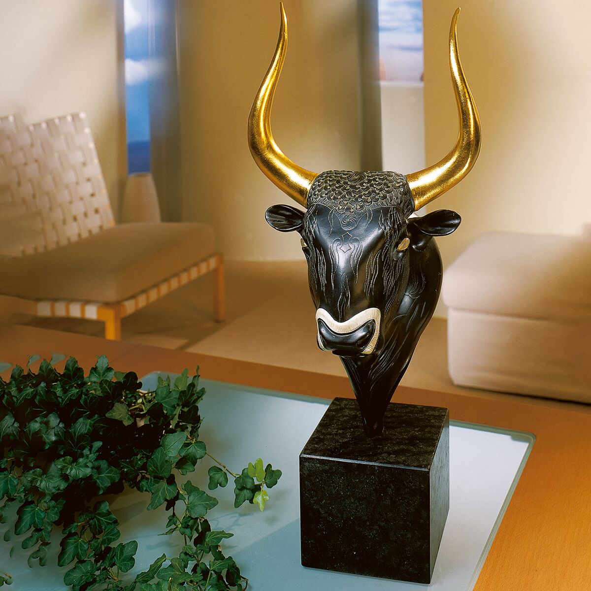 Die Abbildung zeigt eine detailreiche Skulptur eines minoischen Stierkopfs in Grün-Schwarz mit goldenen Hörnern auf einem schwarzen Sockel. Die Szene spielt in einem hellen, modern eingerichteten Wohnzimmer mit beigefarbenen Möbeln und grüner Pflanzen-Deko.