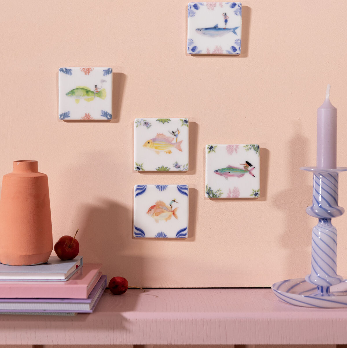 Fünf quadratische Keramikfliesen mit verschiedenen Tier- und Blumenmotiven, die an einer beigefarbenen Wand hängen. Vor der Wand stehen ein Kerzenhalter mit einer lila-weiß gestreiften Kerze und ein korallroter Keramikbehälter auf einem Regal.