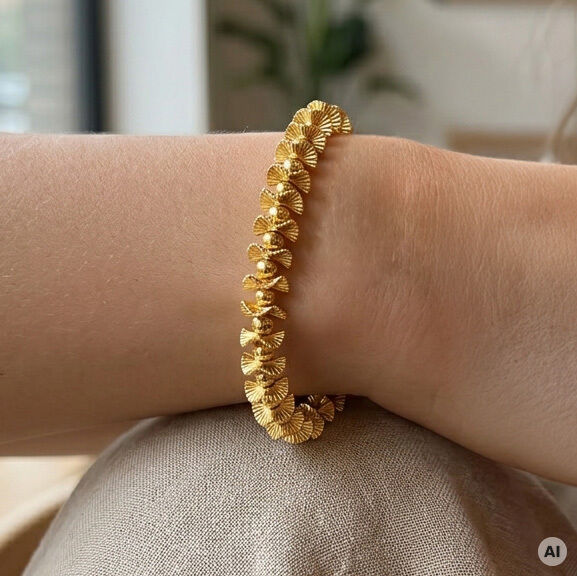 Das Bild zeigt ein elegantes Armband aus 24 Karat vergoldetem Messing von Petra Waszak, Edition Ars mundi, auf einem Handgelenk. Die Glieder des Schmuckstücks sind fächerartig gestaltet und glänzen goldfarben. Im Hintergrund sind unscharfe Wohnraumelemente erkennbar.