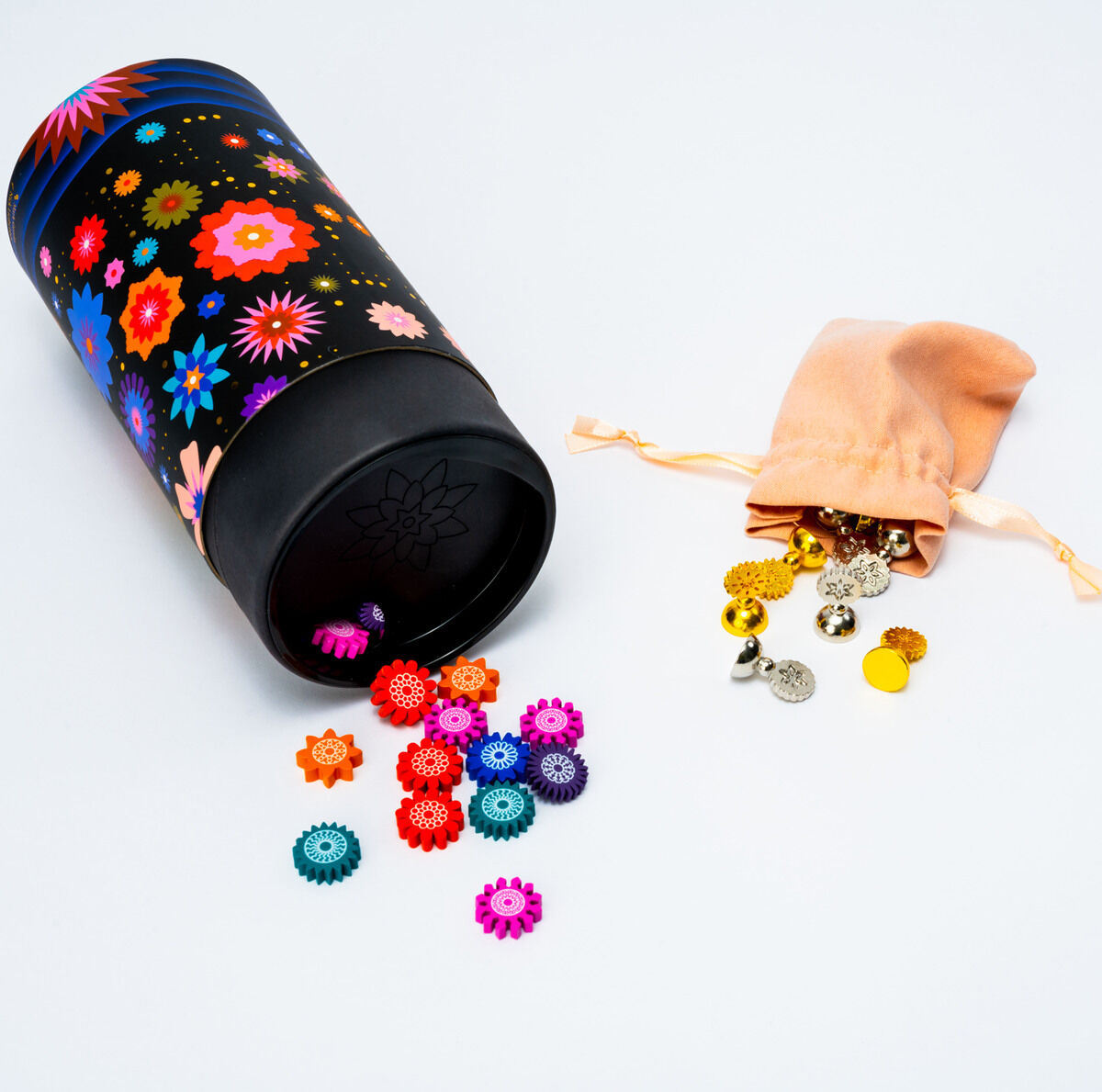 Eine schwarze Aufbewahrungsdose mit buntem Blumenmuster liegt geöffnet auf einer weißen Oberfläche. Daneben befindet sich ein beige Samtbeutel, aus dem goldene und bunte Kunststoffteile herausgefallen sind. Die Szene wirkt verspielt und farbenfroh.