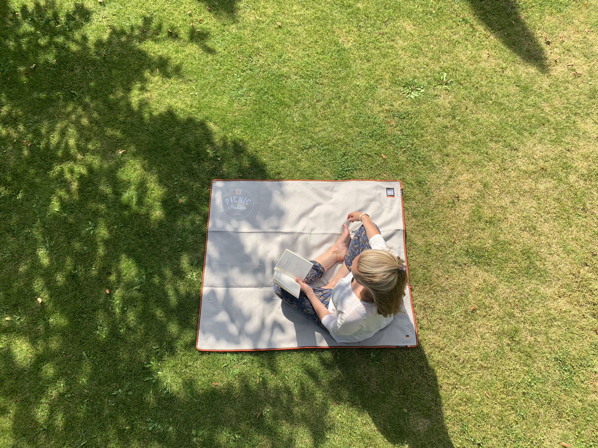 Eine Person sitzt auf einer beige Picknickdecke »Squarepark 11« auf einer grünen Rasenfläche und liest ein Buch, umgeben von natürlichen Schatten.