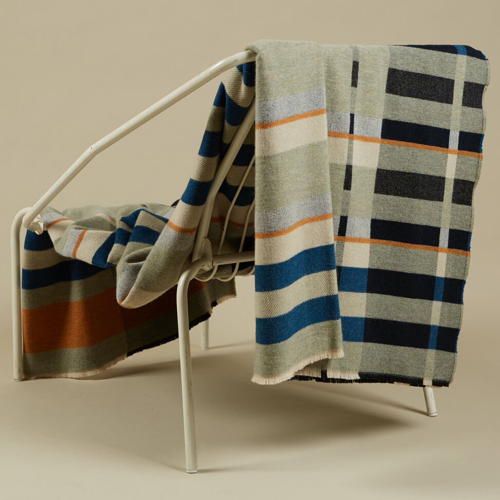 Eine Decke im Bauhaus-Stil, drapiert über einen weißen Metallstuhl. Die Decke hat geometrische Muster in Blau, Orange, Grau, Beige und Schwarz.