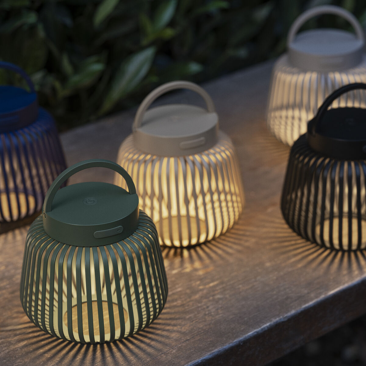 Fünf LED-Leuchten "LITO" von blomus stehen auf einer Steinplatte im Garten. Die Laternen sind gitterförmig gestaltet in den Farben Schwarz, Olivgrün, Creme, Beige und Dunkelblau. Sie verbreiten warmes Licht und werfen dekorative Schatten auf die Oberfläche.