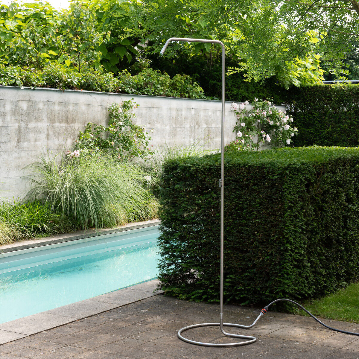 Die Gartendusche »Serpentine« von Weltevree steht auf einer Terrasse neben einem Pool. Sie ist aus silbernem, rostfreiem Edelstahl gefertigt, hat eine schlanke, skulpturale Form und einen runden Standfuß. Der Hintergrund zeigt dichte Hecken und zahlreiche grüne Pflanzen.
