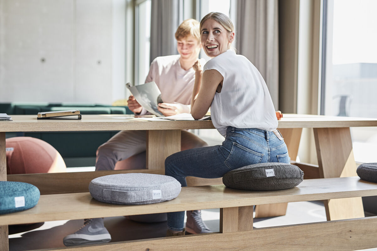 Zwei junge Erwachsene sitzen auf VLUV-Balancekissen »Pil & Ped Stov« an einem langen Holztisch in einem modernen, hellen Wohnzimmer. Im Vordergrund sind mehrere Balancekissen in verschiedenen Farben auf dem Tisch platziert. Die Personen wirken entspannt und glücklich und die Umgebung setzt Komfort und Stil in Szene.
