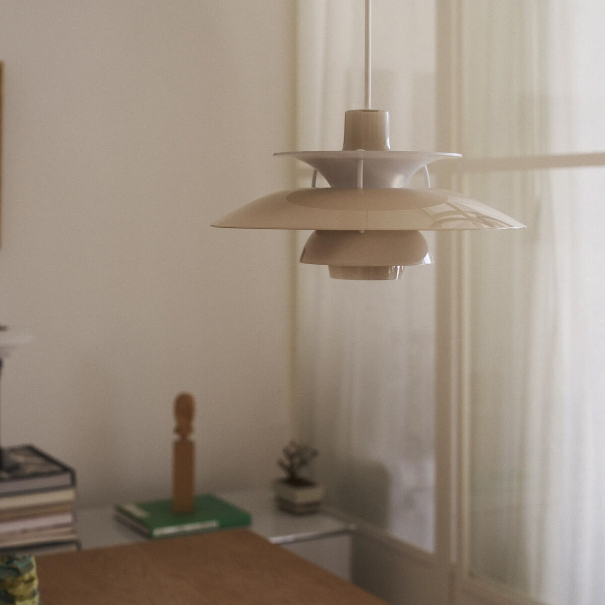 Im Mittelpunkt des Bildes hängt die Hängeleuchte »PH 5 320« von Louis Poulsen in opal beige über einem Holztisch. Die Pendelleuchte besitzt glänzende, kreisförmig angeordnete Schirme aus pulverbeschichtetem Polymer. Im Hintergrund moderne, helle Einrichtung mit Fenstern und Vorhängen.