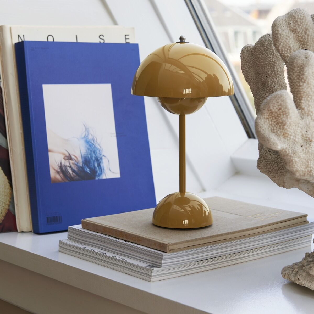 Eine gelbe Flowerpot VP9 Tischleuchte steht auf einem Stapel Bücher auf einer weißen Fensterbank neben blauen Büchern und einer Dekokoralle.