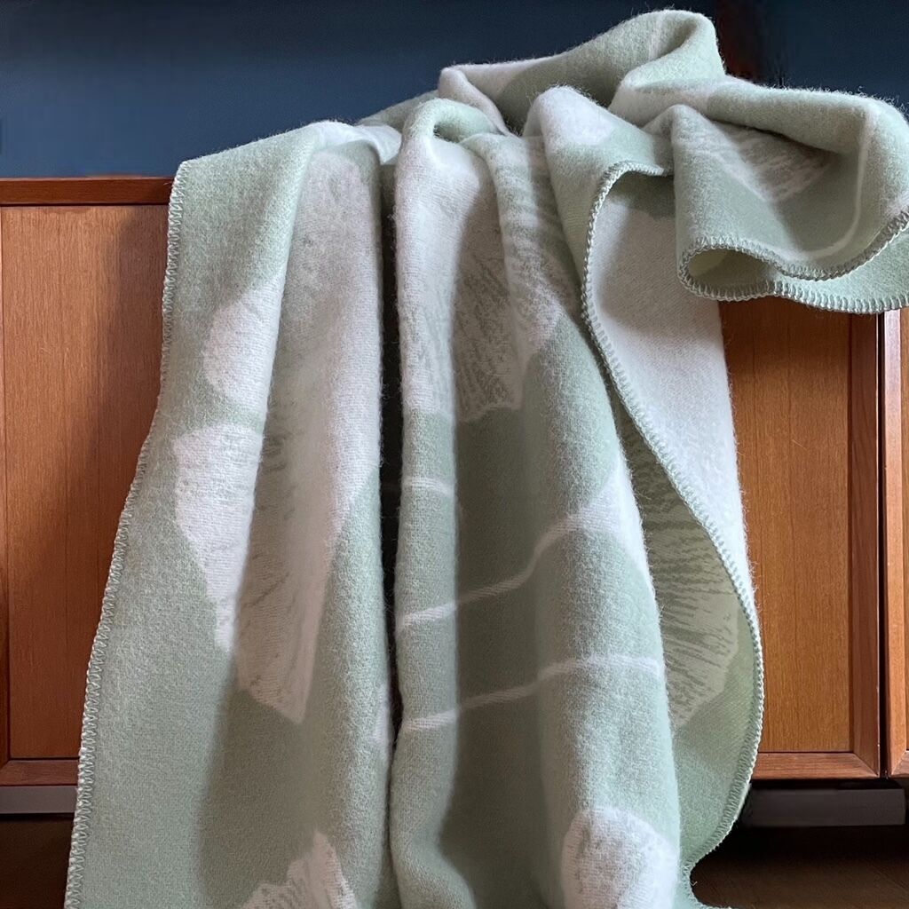 Weiche Decke in Grün- und Weißtönen mit Ginkgo-Muster, drapiert über einem Holzmöbelstück.